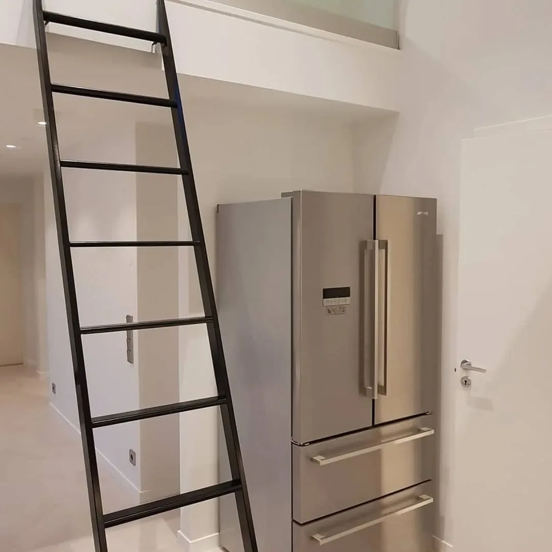 Detalle de acceso al altillo mediante escalera metálica negra junto a frigorífico integrado en reforma de ático