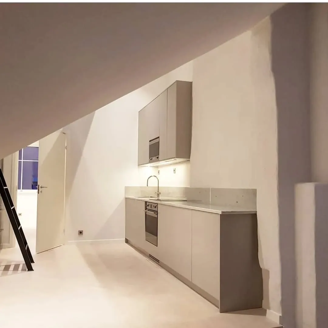 Cocina compacta bajo cubierta inclinada con mobiliario claro, horno integrado y encimera continua
