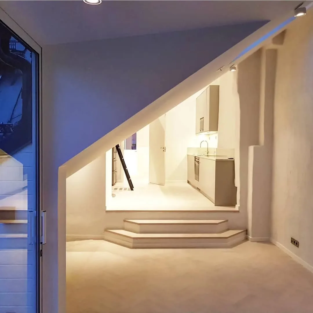 Salón abierto con cocina integrada bajo cubierta inclinada, iluminación cálida y escalones a medida